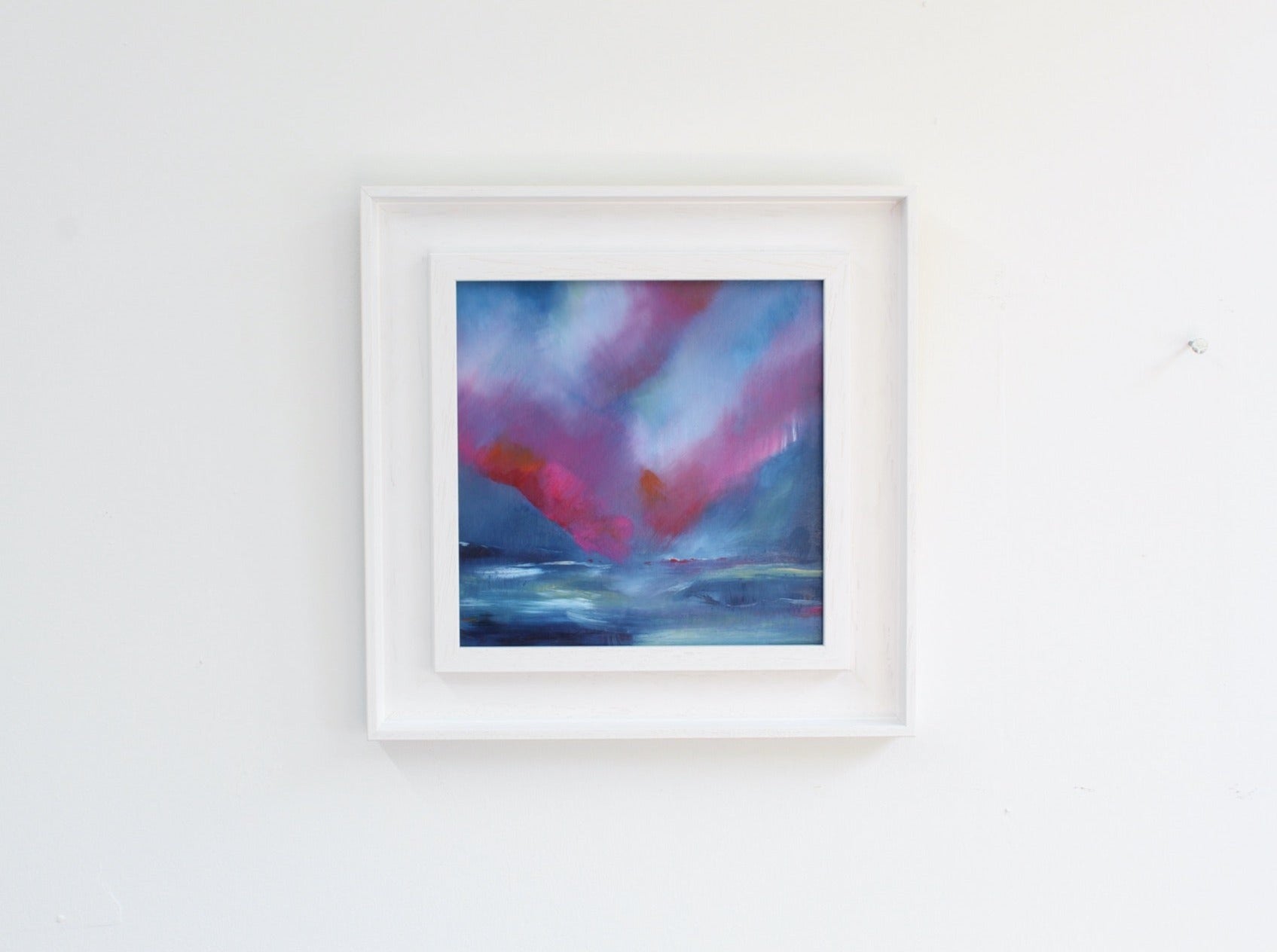 A framed abstract painting with pink highlights on a white wall.